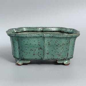 May include: A rectangular ceramic planter with a scalloped rim and four small feet. The exterior features a mottled teal green glaze with dark brown speckles throughout, creating a textured appearance. The vessel has a wide opening and a deep basin.