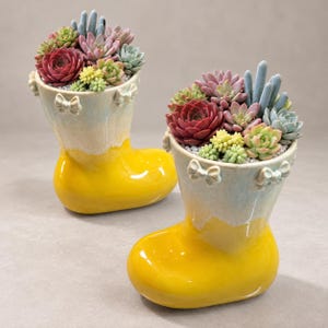 Op de afbeelding: Twee keramische plantenbakken in de vorm van laarzen met een glanzende gele basis en een lichtblauw bovengedeelte. Elke laars is voorzien van kleine decoratieve strikjes langs de rand en gevuld met diverse kleurrijke vetplanten in rood, groen en blauw.