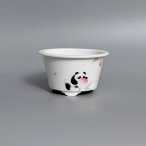 Puede incluir: Un cuenco de cerámica blanca con una ilustración pintada a mano de un oso panda en blanco y negro sosteniendo un melocotón rosa. El cuenco tiene un borde ancho, un cuerpo cónico y pequeños pies en la base, decorado con delicadas hojas verdes y flores rosas.