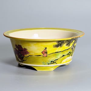 May include: A yellow ceramic bowl with a wide rim and a cream-colored interior. The exterior features a hand-painted landscape scene with a figure in a red robe, a red-leafed tree, and black ink-style pine trees on a yellow background.