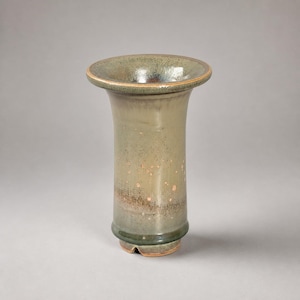 以下が含まれることがあります： 広いフレア状の縁と円筒形の胴体を持つ陶器製の花瓶。表面はオリーブグリーンと落ち着いた茶色の光沢のある釉薬で仕上げられており、繊細な斑点模様と、切り込みのある底部の近くには暗い質感の帯が見られます。
