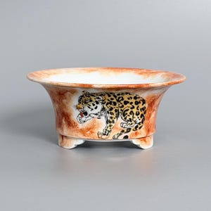 May include: A ceramic bowl with a wide, flared rim and three small feet. The exterior features a hand-painted leopard with black spots on a tan coat, set against a mottled, burnt orange and white background. The interior is a plain white finish.