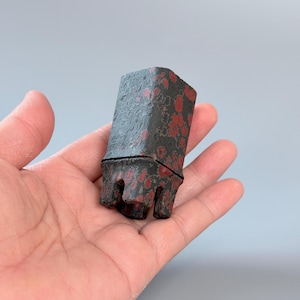 Puede incluir: Un objeto pequeño, rectangular y texturizado con un color base gris carbón oscuro y manchas rojas irregulares y moteadas en su superficie. El objeto presenta una costura horizontal distintiva y una base con cuatro puntas dentadas hacia abajo.