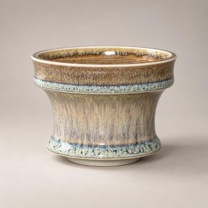 May include: A ceramic planter with a flared rim and a tapered base. The exterior features a reactive glaze with vertical drip patterns in shades of tan, beige, and brown, accented by a band of mottled light blue and dark blue near the rim and base.