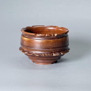 Puede incluir: Un cuenco de cerámica de color marrón oscuro con un acabado esmaltado y liso. El cuenco presenta un borde central horizontal ancho y un borde decorativo dentado o festoneado tanto en la parte superior como en la sección media, creando una apariencia escalonada.