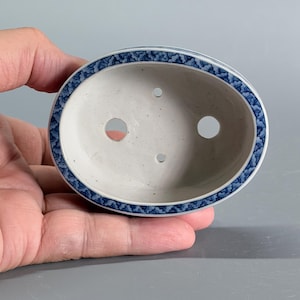 May include: An oval ceramic bonsai pot with a white interior and a blue and white cloud-like pattern along the rim. The base of the pot features two large drainage holes and two smaller holes, providing a clear view of the interior structure.