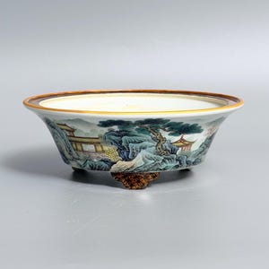 May include: A shallow ceramic bowl with a wide rim and three small feet. The exterior features a detailed landscape painting with green trees, blue rock formations, and yellow buildings. The rim is accented with a thin brown and gold border.
