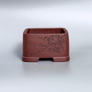 May include: A rectangular, dark reddish-brown clay planter with a matte, textured finish. The front side features a detailed, dark engraved illustration of a small bird perched on a branch among flowers and foliage, with four small feet.