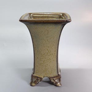 Puede incluir: Un jarrón de cerámica de borde cuadrado con silueta acampanada y esmalte moteado en beige y marrón. El recipiente presenta una abertura ancha, un cuerpo cónico y se apoya sobre cuatro pies distintos y ornamentados con acabado marrón oscuro.