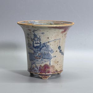 May include: A ceramic planter with a flared rim and three small feet, featuring a speckled beige glaze. The exterior is decorated with a blue ink illustration of a bearded figure in traditional armour riding a horse, accented by reddish-brown patches.