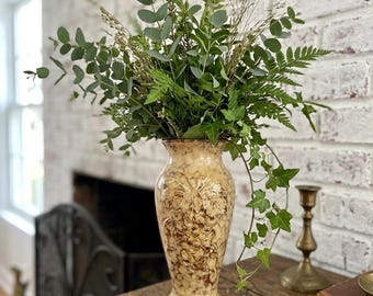 May include: A tall, beige vase with a grape design holds a lush arrangement of green foliage. The vase sits on a wooden surface, with a white brick wall in the background. A brass candlestick and a book are also visible.