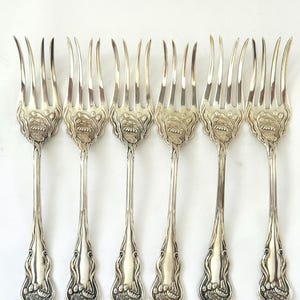 May include: A set of six silver forks with ornate floral designs on the handles and tines. The forks are arranged in a row against a white background, showcasing their elegant details. The tines are long and slender, and the handles have a detailed pattern.