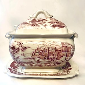 May include: A cream-colored ceramic tureen with a matching lid and rectangular base plate. The piece features a detailed maroon transferware pattern depicting a castle, a bridge, and a small boat on a river, with ornate handles on the sides.