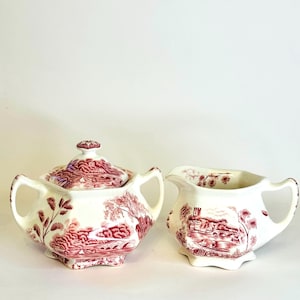 May include: A cream-colored ceramic sugar bowl with a lid and a matching creamer pitcher, both featuring a detailed maroon transferware landscape pattern depicting a river, a stone bridge, and trees with a scalloped base and side handles.