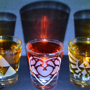 May include: Three shot glasses filled with amber and red liquid. The glasses have white etched designs: a triangle, a heart, and a stylized emblem. Shadows of the glasses are cast on the background.