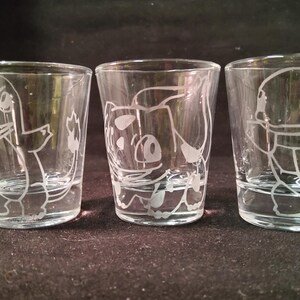May include: Three clear shot glasses with etched cartoon character designs. Each glass features a different character, with detailed white outlines. The glasses are cylindrical with a slight taper, and are displayed against a dark background.