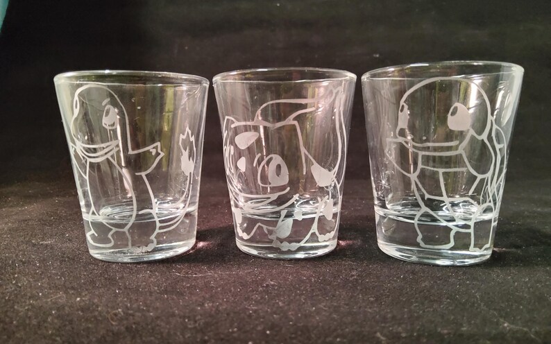May include: Three clear shot glasses with etched cartoon character designs. Each glass features a different character, with detailed white outlines. The glasses are cylindrical with a slight taper, and are displayed against a dark background.