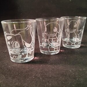May include: Three clear shot glasses with etched cartoon character designs. The glasses are cylindrical with a flared rim. The etched designs are white and feature different characters.