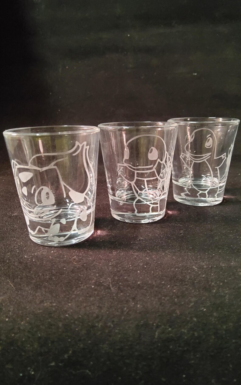 May include: Three clear shot glasses with etched cartoon character designs. The glasses are cylindrical with a flared rim. The etched designs are white and feature different characters.