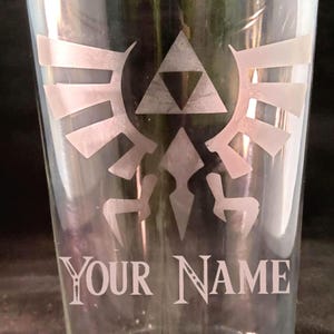 May include: A clear glass pint glass with a frosted design of the Triforce symbol from the Legend of Zelda video game series. The text "YOUR NAME" is also frosted on the glass.