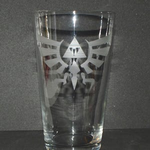 May include: A clear glass pint glass with a white etched design of the Triforce symbol from the Legend of Zelda video game series.