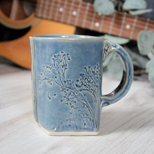 May include: A blue ceramic mug with a white floral design. The mug has a slightly irregular shape and a handle.