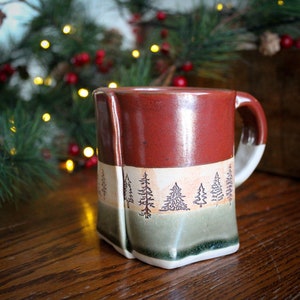 May include: A red, white, and green ceramic mug with a design of pine trees. The mug has a slightly rounded shape and a handle.