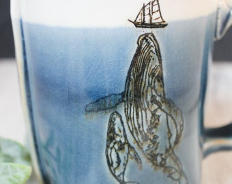 May include: A blue ceramic mug with a whale and a sailboat design. The whale is in the center of the mug and the sailboat is on top of the whale. The mug has a crackled glaze.