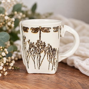 May include: A cream-colored ceramic mug featuring a dark brown, etched-style design of dragonflies hovering above tall, thin meadow grasses. The mug has a thick, hand-formed handle and a slightly irregular, organic shape with a smooth finish.