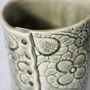 May include: Close-up of a ceramic mug with a floral lace pattern. The mug is a pale green color with darker green accents in the floral design. The mug has a unique, textured surface and a button-like detail.