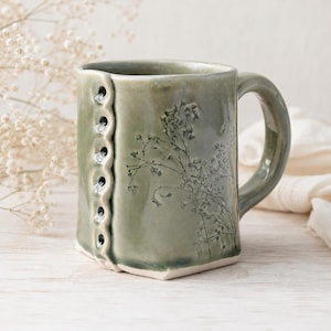 May include: A sage green ceramic mug featuring a vertical row of seven small, dark, circular indentations along the left side and a delicate, dark botanical print of wildflowers across the front surface, complete with a curved handle on the right.
