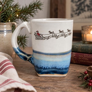May include: A ceramic mug with a white upper section featuring a black line drawing of Santa Claus in a sleigh pulled by reindeer. The lower portion of the mug has a reactive glaze finish in shades of blue and cream, transitioning to a dark blue base.