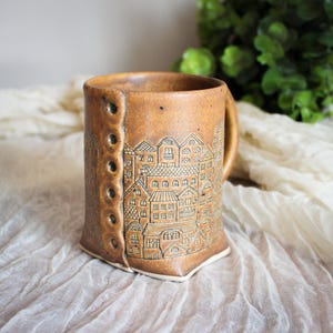May include: A cylindrical ceramic mug with a speckled amber-brown glaze. The exterior features a vertical row of six circular indentations resembling buttons and a detailed, etched illustration of a dense cityscape with multiple buildings.