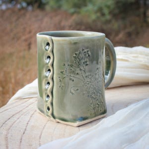 May include: A handmade, sage green ceramic mug with a square shape and a handle. The mug features a floral design and a decorative, wavy edge with small holes. The mug is displayed on a wooden surface.