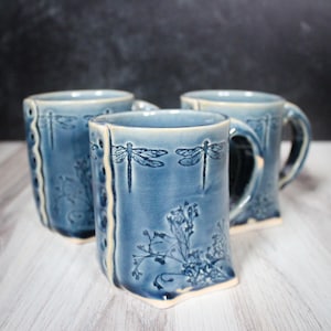 May include: Three ceramic mugs with a glossy blue glaze, featuring embossed dragonfly and floral patterns. Each mug has a distinct vertical side panel with a row of small circular indentations and a light-colored base rim.