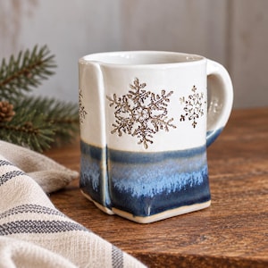 May include: A ceramic mug with a square-shaped base and a rounded handle. The upper portion is off-white with embossed brown snowflake patterns, while the lower portion features a gradient of blue and dark navy glaze with a textured finish.