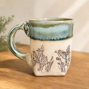 May include: A ceramic mug with a cream-colored base featuring black line art of birds and wildflowers. The upper portion and the handle are finished in a glossy, mottled teal and dark green glaze, creating a distinct contrast with the lower body.