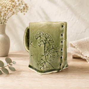 May include: A sage green ceramic mug featuring a dark, etched ginkgo leaf botanical illustration on the front. The mug has a unique, slightly squared shape with a vertical row of small, dark circular indentations along the right side edge.