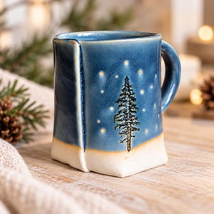 May include: A handmade ceramic mug with a square-shaped base and a rounded handle. The exterior features a glossy blue glaze on the upper portion with a painted black pine tree and yellow stars, transitioning to a matte white base at the bottom.