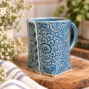 May include: A ceramic mug with a glossy blue glaze and a textured surface featuring an embossed floral and lace-like pattern. The mug has a vertical seam detail along the front with small, dark, circular indentations resembling buttons.