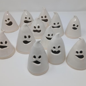 May include: A collection of twelve small, white, cone-shaped ceramic ghost figurines. Each ghost features a smooth, glossy finish with cut-out eyes and a mouth, creating a hollow opening. The figures are arranged in a group on a plain surface.