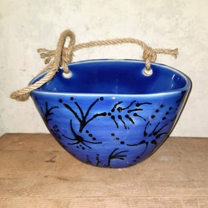 May include: A vibrant blue ceramic hanging planter with a rope handle. The planter features black abstract floral patterns. The planter is displayed on a wooden surface. The planter is approximately 20 cm in diameter.