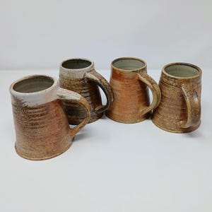 May include: Four handmade ceramic mugs with large handles. The mugs have a rustic, textured appearance with shades of brown, tan, and cream. The mugs are cylindrical with a slightly flared top, perfect for enjoying a beverage.