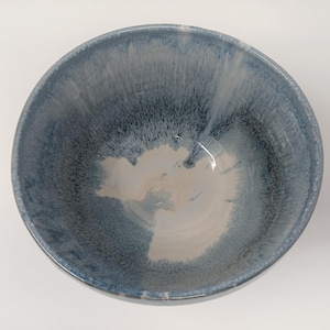 Op de afbeelding: Een handgemaakte keramische kom met een glanzende, getextureerde afwerking. De kom heeft een mix van blauwe en witte tinten, waardoor een uniek marmer effect ontstaat. De binnenkant is lichtbeige. Geschikt voor het serveren van voedsel.