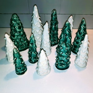 May include: A collection of ceramic Christmas tree ornaments in white and green. The trees are all different sizes and shapes, and some have a textured finish.