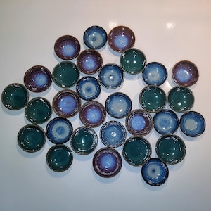 Puede incluir: Una colección de 24 cuencos pequeños y redondos de cerámica en varios tonos de azul, verde y marrón. Los cuencos tienen un esmalte único y moteado y están dispuestos en un patrón suelto y orgánico.