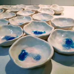 May include: A collection of small, white ceramic bowls with blue and white glaze. The bowls have a wavy, organic shape and are arranged in a staggered pattern.