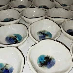 Puede incluir: Una colección de pequeños cuencos de cerámica blanca con esmalte azul y verde. Los cuencos tienen una forma única, texturizada y ligeramente irregular.