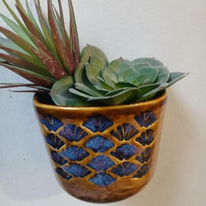 Puede incluir: Una maceta de pared de cerámica marrón con un diseño de abanico azul y negro, que contiene una suculenta verde y una planta roja y verde. La maceta está montada en una pared blanca, mostrando una estética decorativa y natural.
