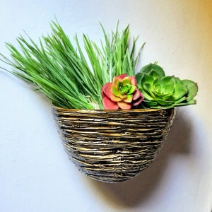 Op de afbeelding: Een gouden, getextureerde keramische pot met een groene succulent en een roze-groene succulent. De pot is gemonteerd op een witte muur.